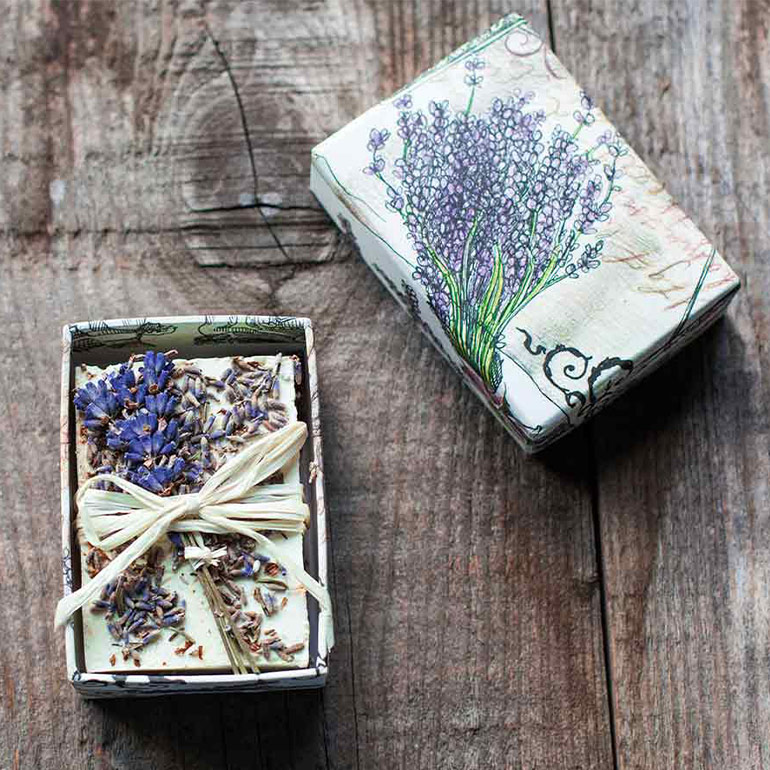 A gift box with the lid removed showing a bar of lavender soap within it, there is string tied around the soap and lavender flowers within the box too.