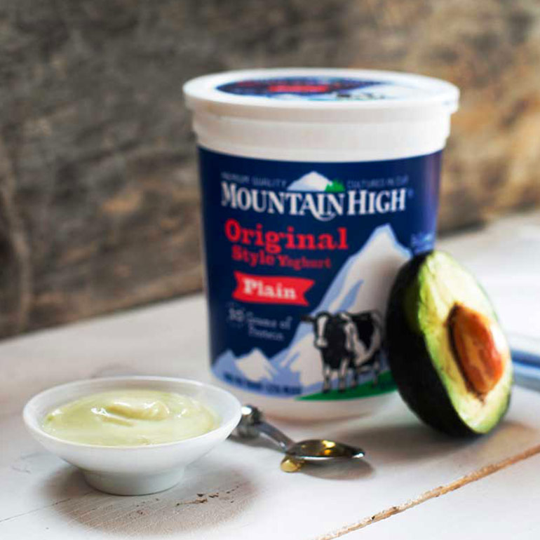 A milk mask in a bowl in front of an unopened pot of Mountain High Plain Yoghurt, half an avocado is resting against the pot and a teaspoon sits in front.