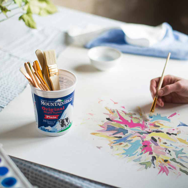 A pot of Mountain High Plain Yoghurt being used as a pot for paint brushes whilst someone is halfway through painting a watercolour next to the pot.